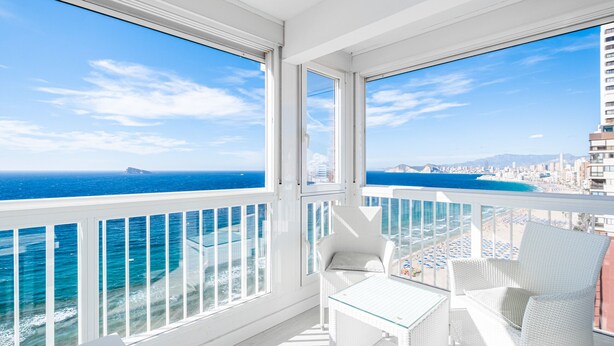 A balcony with white wicker furniture, a glass table, and a view of the ocean and cityscape. - Veracruz 18-A (Benidorm)