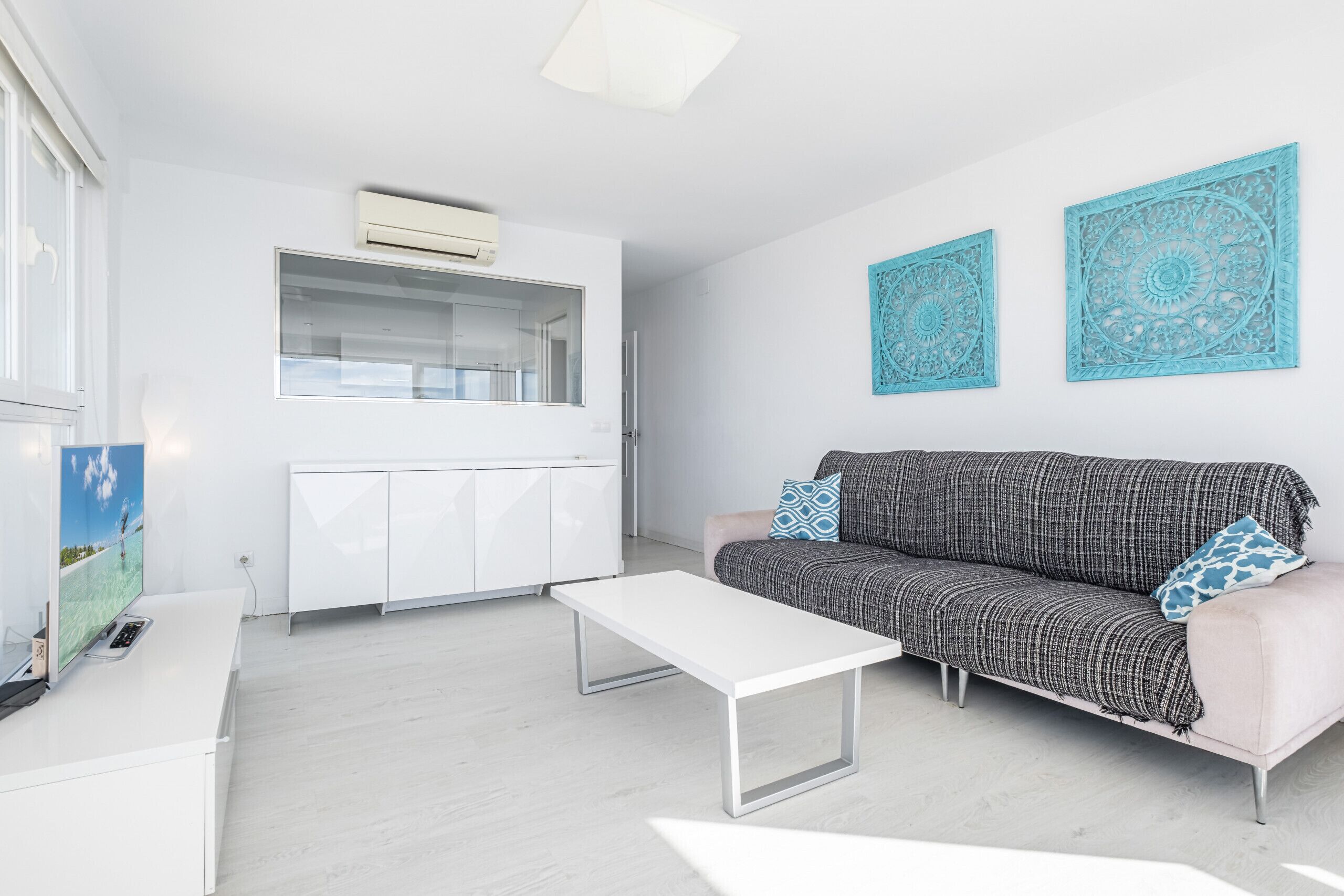 A modern living room with a white sofa, a white coffee table, and a TV on a white cabinet. There are two blue decorative wall pieces above the sofa.