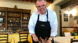 Eine Person in weißem Hemd und schwarzer Schürze bereitet Essen in einer Küche mit Holzmöbeln zu, im Hintergrund ist ein Weinkühlschrank zu sehen.
