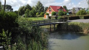 Une maison avec un toit rouge, un pont en bois enjambant un étang, et une végétation luxuriante tout autour.