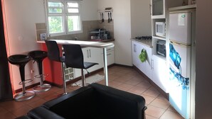 Een kleine keuken met een wit aanrecht, twee zwarte barstoelen, een eettafeltje, een koelkast en een gasfornuis.