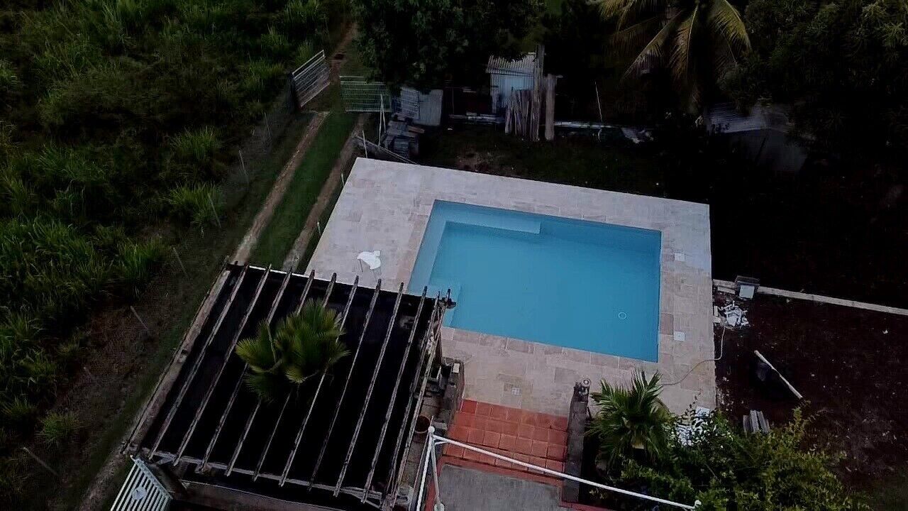 Luchtbeeld van een rechthoekig zwembad, omringd door een stenen terras, met een metalen dakconstructie aan de linkerkant en palmbomen op de achtergrond.