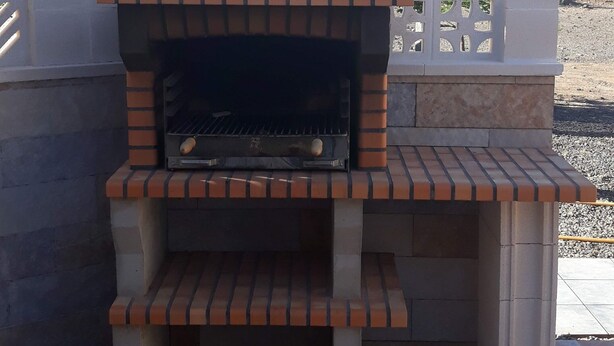 An outdoor brick oven barbecue area with a chimney, surrounded by a white fence and a tiled patio.