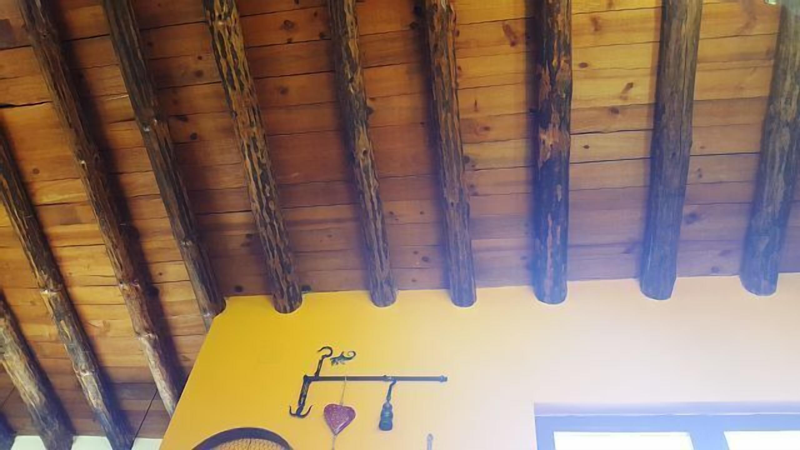 Techo de madera con vigas a la vista y una pared amarilla con una lámpara colgante sencilla.