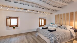 A bedroom with a large bed, two windows, wooden ceiling beams, and string lights.
