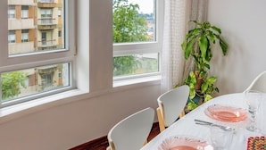 Zona pranzo con tavolo apparecchiato per due, ampie finestre, una pianta in vaso e vista su un edificio esterno.