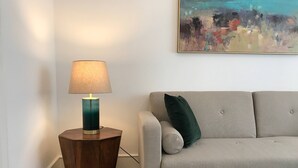 A living room with a round wooden side table, a lamp, a grey sofa, a green pillow, and a painting on the wall.