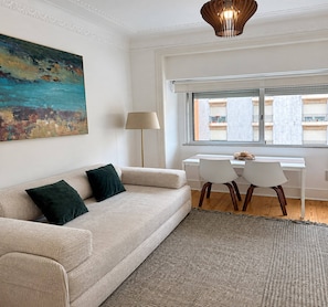 A modern living room with a sofa, a dining table with chairs, a lamp, and a painting on the wall.