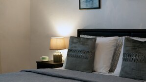 A neatly made bed with a grey blanket, two pillows with a geometric pattern, a bedside table with a lamp, and a framed picture on the wall.