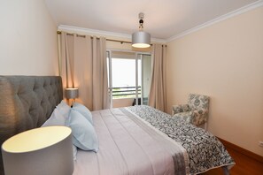 Een slaapkamer met een bed, nachtkastje, stoel en raam met gordijnen.