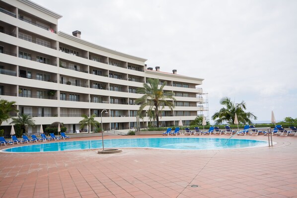 Een hotel met een zwembad, omringd door blauwe ligbedden en palmbomen.