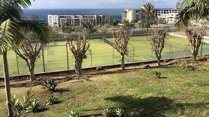 Una cancha de tenis rodeada de palmeras y edificios, con vista al océano al fondo.
