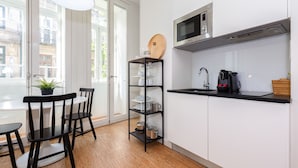 A modern kitchen with white cabinets, a black countertop, a microwave, and a coffee maker. There is a wooden shelf with dishes and a potted plant on the windowsill.