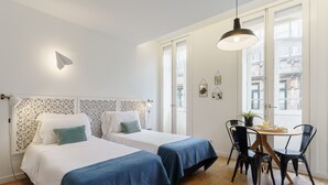 A hotel room with two beds, a dining table with chairs, and a window with a view of buildings.