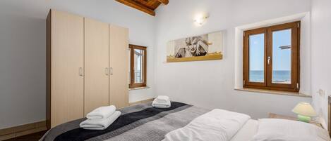 A bedroom with a bed, a wardrobe, a painting, and a window with a view of the sea.