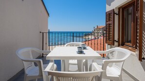 Un balcón con una mesa y sillas blancas, con vistas a una playa y el mar, con edificios al fondo.