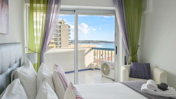A hotel room with a bed, a chair, a fan, and a view of the beach and buildings. - Apartment Rocha | Sea View | Concorde Sky (Portimão)