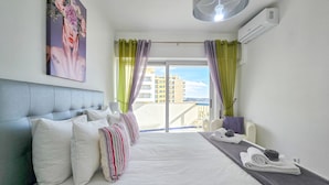 A hotel room with a bed, pillows, a bedside table with towels, a fan, and a painting on the wall. - Apartment Rocha | Sea View | Concorde Sky (Portimão)