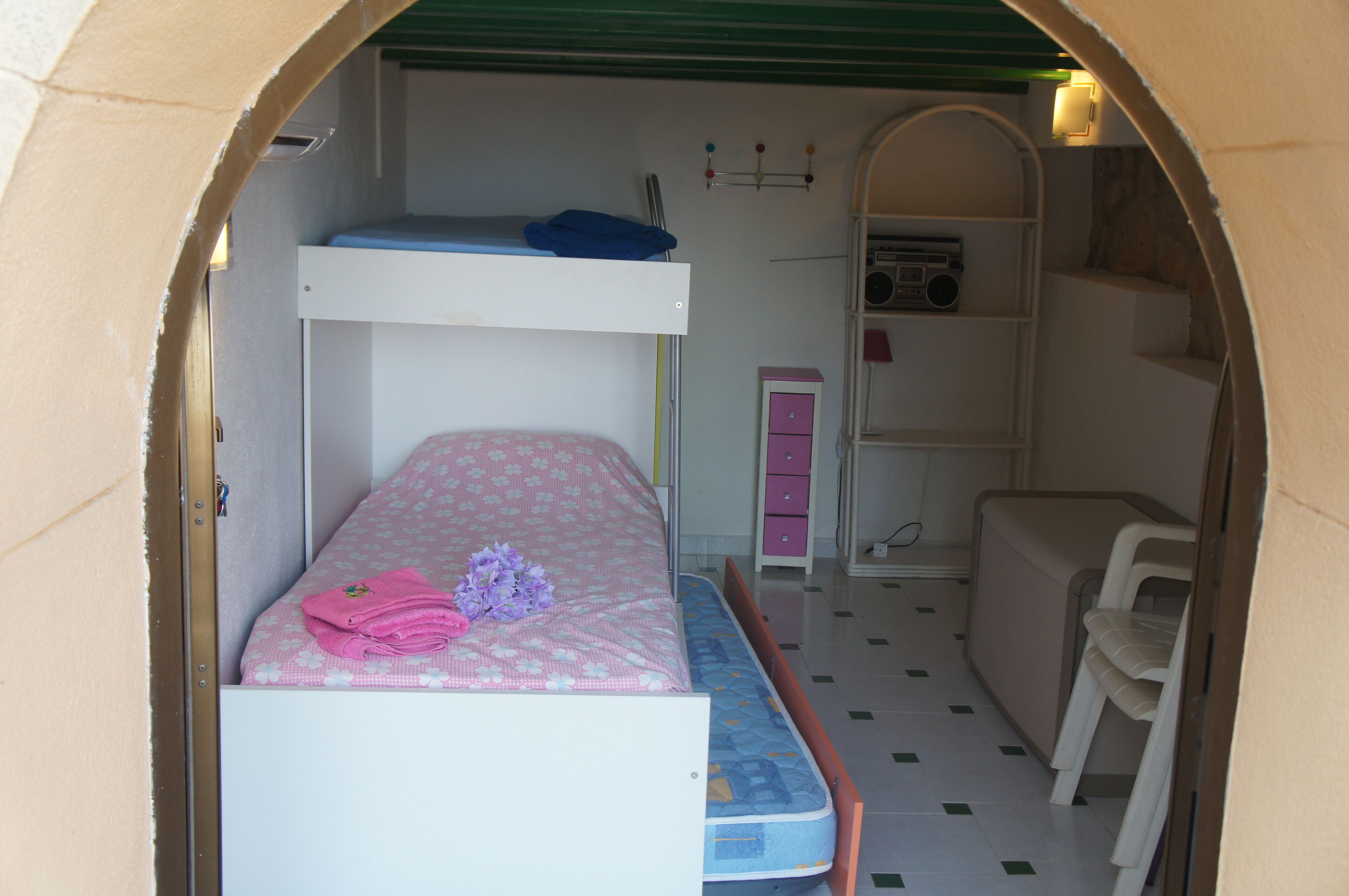 A bunk bed with pink bedding and a blue mattress, a pink dresser, a radio, and a white chair in a room with a tiled floor.