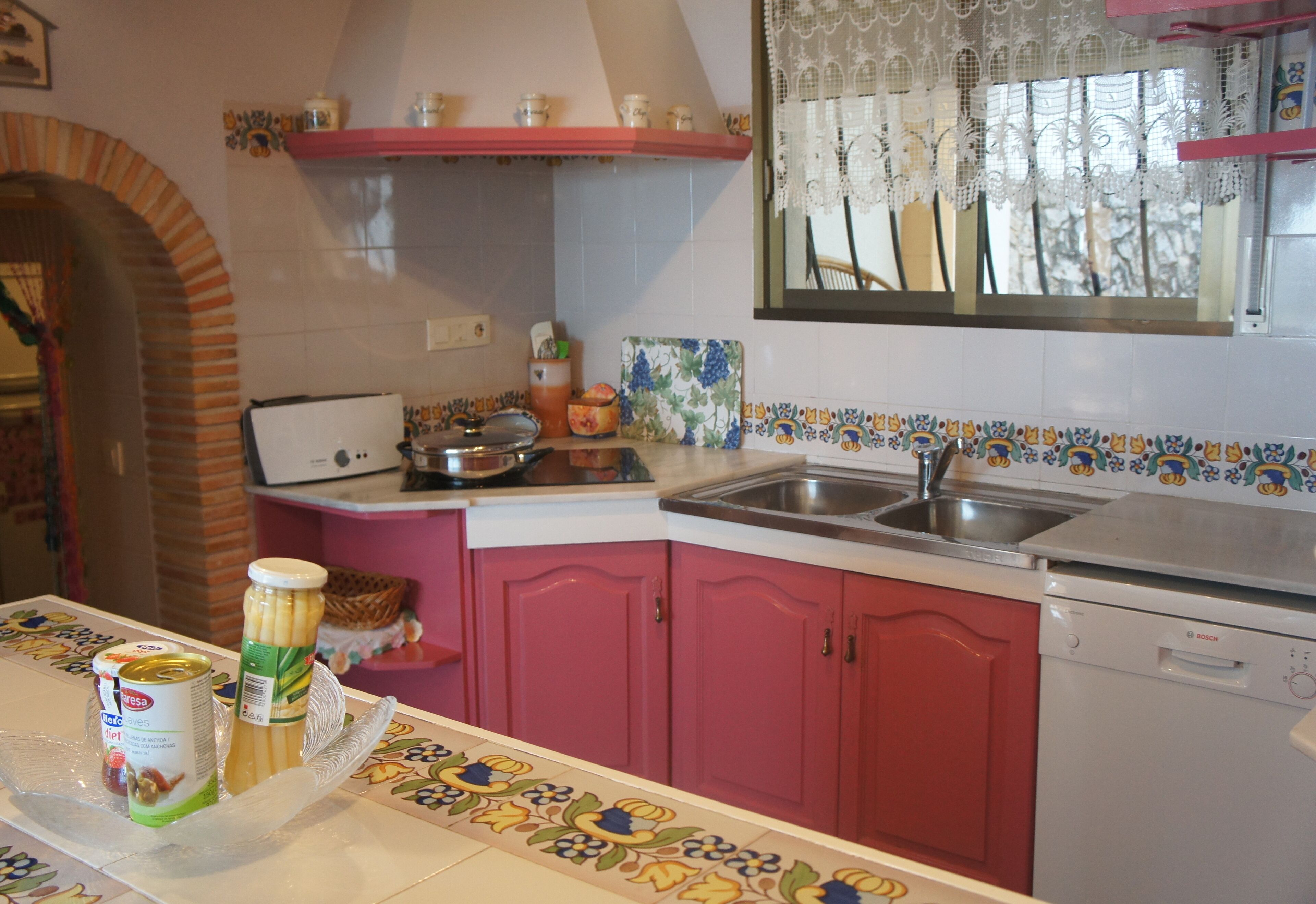 A kitchen with pink cabinets, a Bosch dishwasher, and a window with lace curtains.