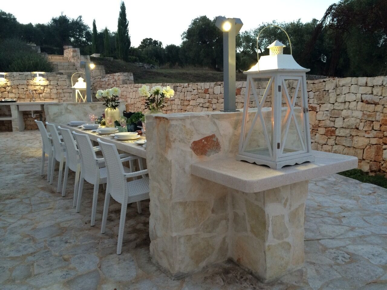 An outdoor dining area with stone walls, white chairs, and a lantern.
