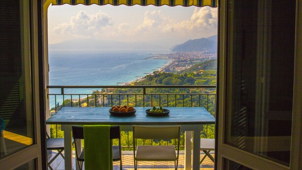Un balcón con mesa y sillas, con vistas al mar, montañas y un paisaje urbano.