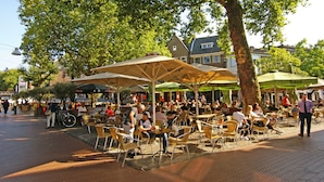 An outdoor café with tables and chairs under umbrellas, surrounded by trees and buildings.