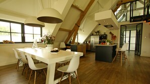 A spacious dining area with a white table and chairs, a kitchen in the background, and a loft space above.