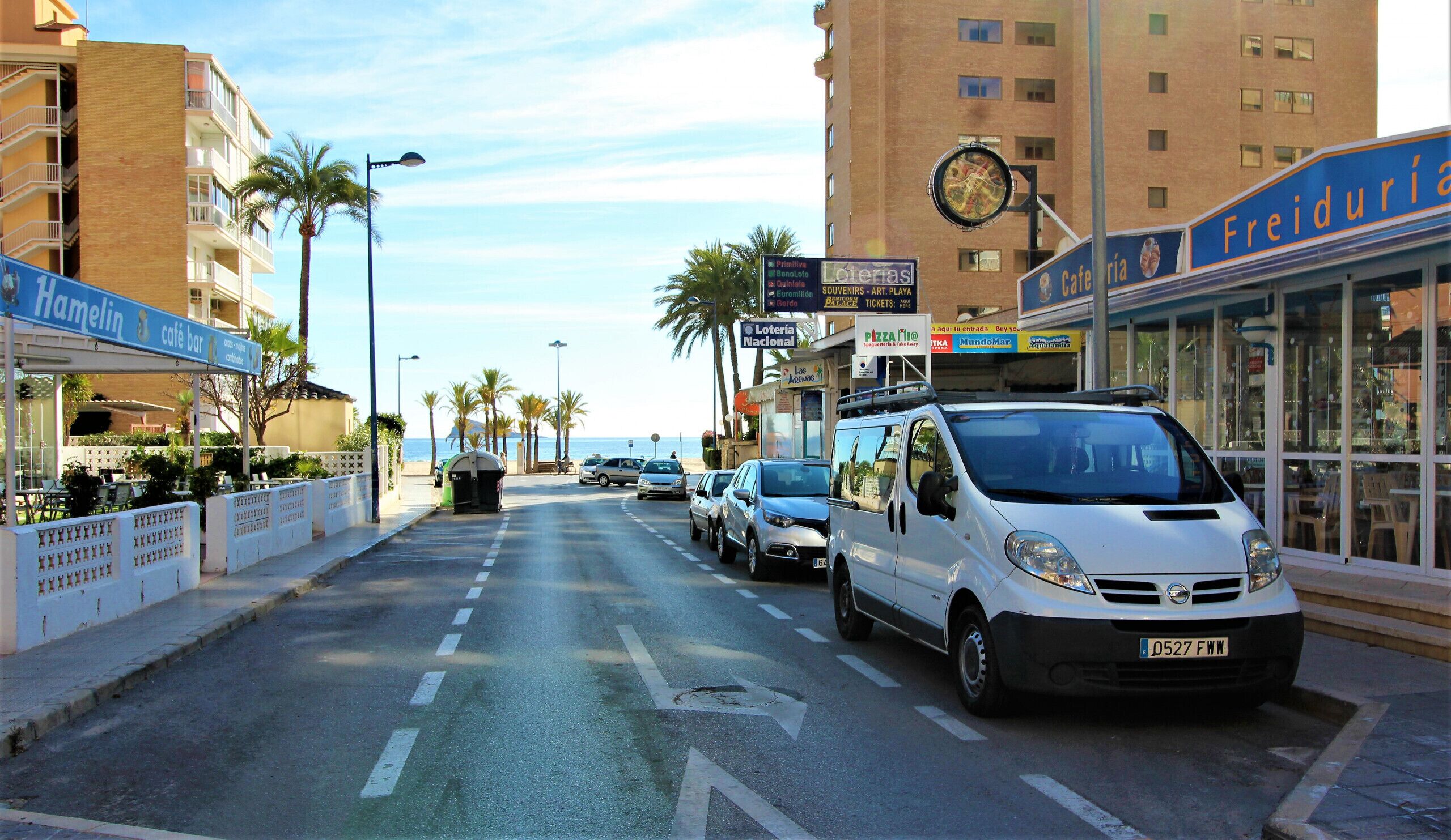 Een straat met geparkeerde auto's, palmbomen en gebouwen met borden voor een café-bar en een restaurant.