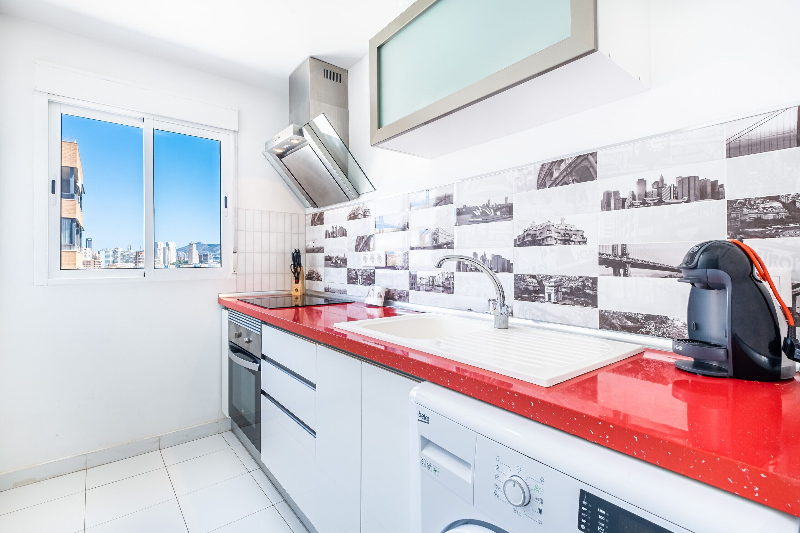 A modern kitchen with a red countertop, a built-in dishwasher, and a large window with a city view.