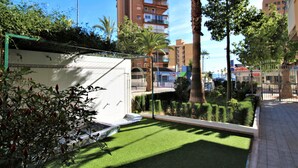 Un patio con césped artificial, un edificio blanco con balcón y un edificio residencial alto al fondo.