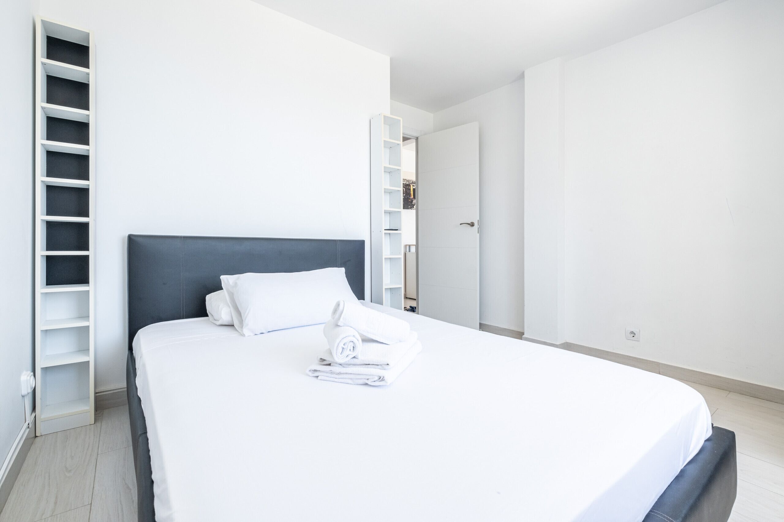 Un lit bien fait, avec du linge de lit blanc, une tête de lit grise et une table de chevet blanche sur laquelle est posée une serviette pliée. Une armoire blanche avec un miroir est visible sur la droite.
