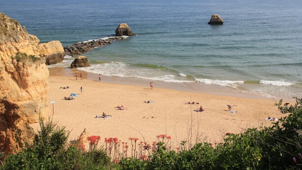 Uma praia com falésias rochosas, mar calmo e pessoas tomando sol.