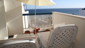 Uma varanda com vista para o mar, cadeiras brancas, um guarda-sol listrado e plantas em vasos.