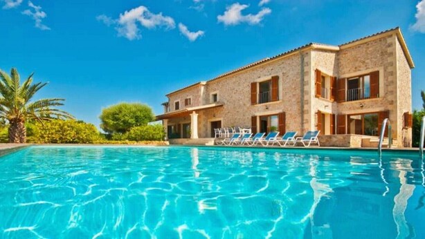 A poolside view of a two-story stone building with a clear blue pool, lounge chairs, and palm trees.