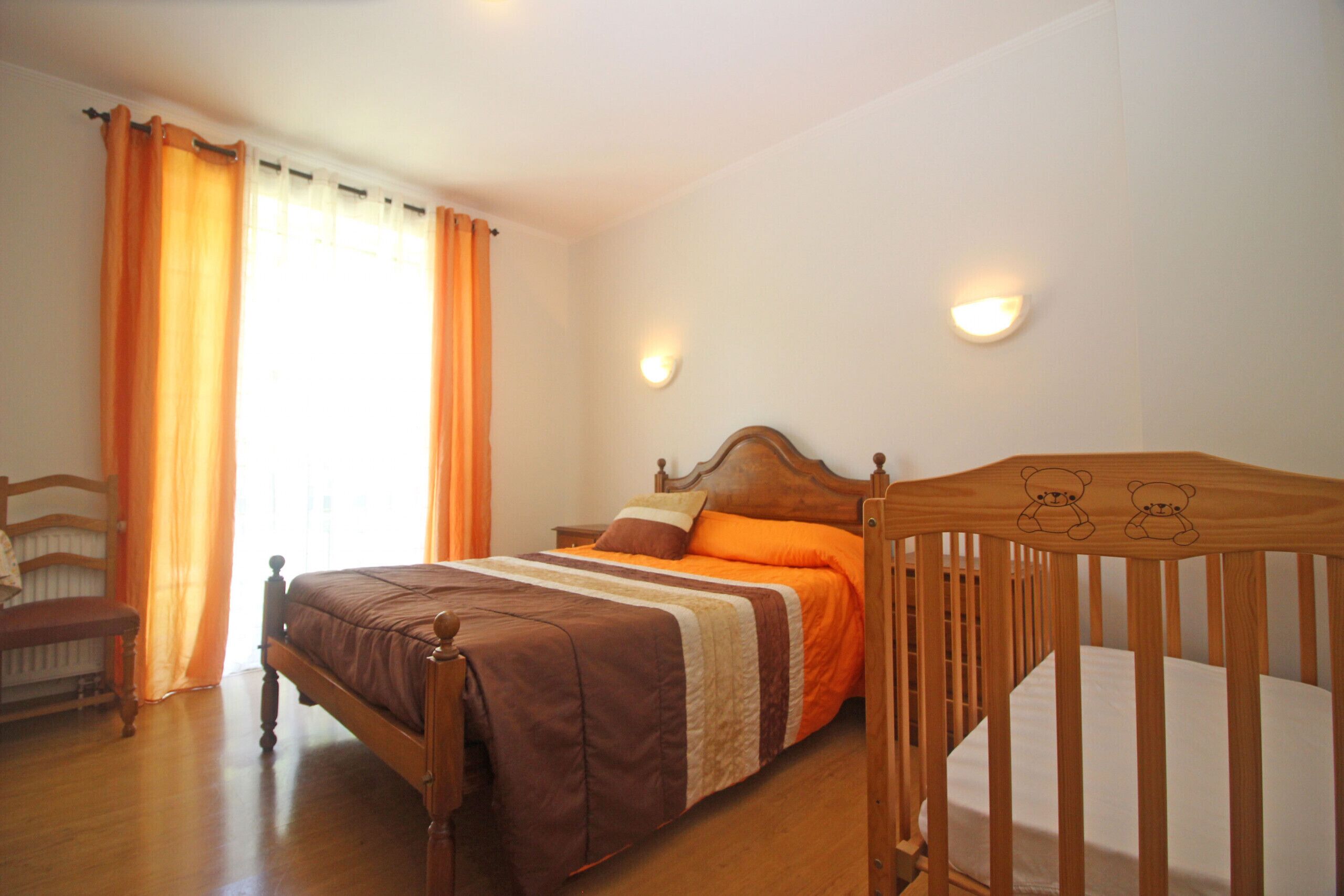 A bedroom with a wooden bed, orange bedding, a wooden crib, and two wall-mounted lights.