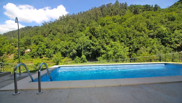 A swimming pool with a metal ladder, surrounded by a concrete deck, set against a backdrop of lush green hills. - House with private pool near Gerês (Terras de Bouro)