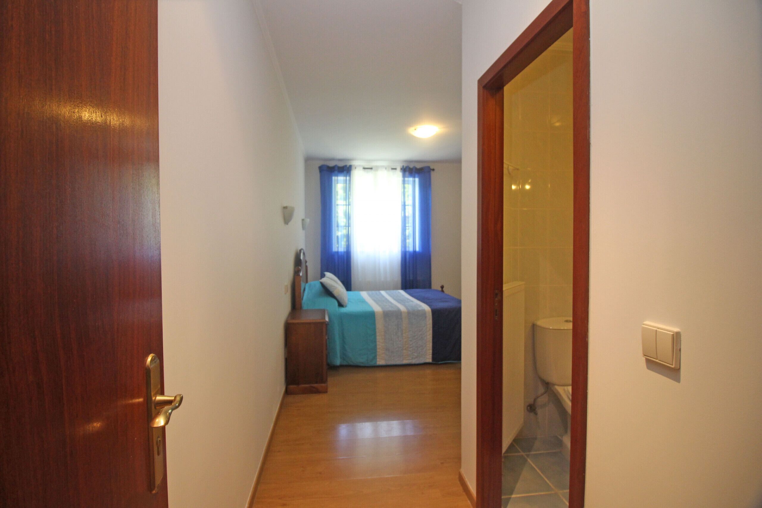 Ein schmaler Flur führt zu einem Schlafzimmer mit einem Bett, einem Nachttisch und einem Fenster mit Vorhängen.