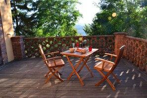 An outdoor patio with a wooden table and two chairs, a brick wall with greenery, and a view of trees and hills.