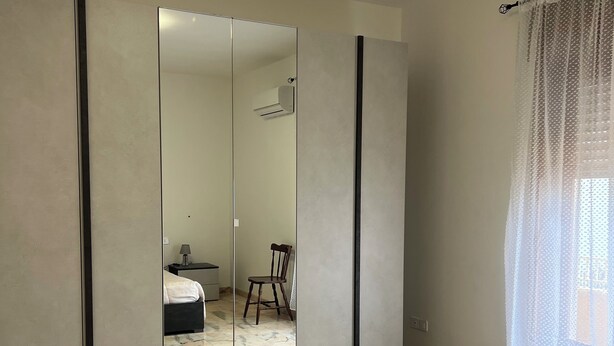 Quarto com um espelho grande, uma cama, uma cadeira e uma janela com cortinas leves.