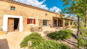 A rustic stone building with wooden doors and shutters, a patio with benches, and a garden with shrubs.