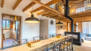 A dining area with a long table, chairs, hanging pendant lights, and a fireplace.