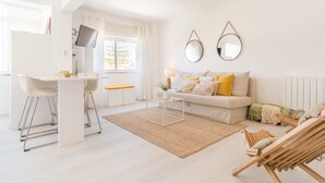 A bright, modern living room with a white sofa, a dining area with a white table and chairs, and a large window providing natural light. - Parede Ocean (Parede)