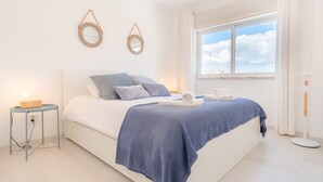 A bedroom with a bed, pillows, a bedside table, a lamp, a window with a view, and two wall-mounted mirrors. - Parede Ocean (Parede)
