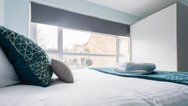 Habitación de hotel con un ventanal grande, una cama con ropa blanca, un plaid verde azulado y una toalla doblada sobre la cama.