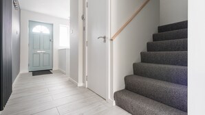 Un pasillo con suelo de madera clara, una puerta azul claro y una escalera con peldaños cubiertos de moqueta gris.
