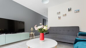 Una sala moderna con un televisor de pantalla plana, una mesa de centro redonda blanca, un sofá gris, cojines color turquesa y un jarrón rojo con flores.