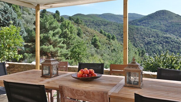 Une table en bois, sur laquelle se trouve un saladier rempli de fruits, deux lanternes et des chaises, le tout placé sous une tonnelle offrant une vue sur la montagne.