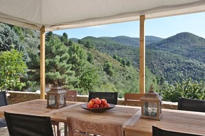 Une table en bois, sur laquelle se trouve un saladier rempli de fruits, deux lanternes et des chaises, le tout placé sous une tonnelle offrant une vue sur la montagne.
