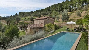 Une piscine bordée de pierres, un bâtiment en pierre avec un toit en tuiles et une zone herbeuse avec des arbres.